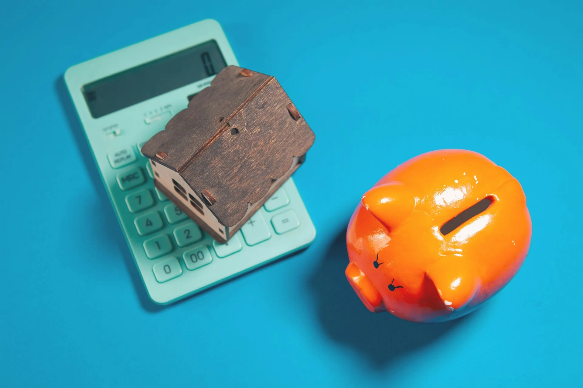 Miniature wooden house on a calculator next to a piggy bank representing mortgage savings